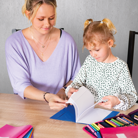Eine Frau und ein kleines Mädchen sitzen an einem Tisch und schauen sich ein Buch an. Bunte Stifte und Notizblätter liegen auf dem Tisch.