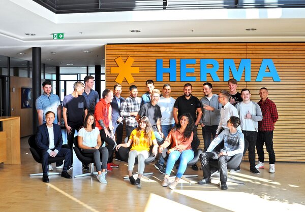 Gruppensitzung von Mitarbeitern vor einer Holzwand mit dem Schriftzug "HERMA". Die Personen stehen und sitzen in verschiedenen Posen.