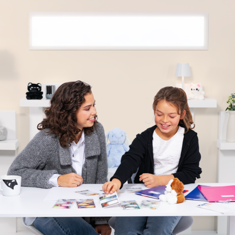 Zwei Mädchen sitzen an einem Tisch, umgeben von Fotos und Notizmaterialien, während sie miteinander sprechen und lachen.