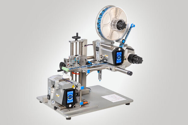 Eine Etikettiermaschine mit verschiedenen Komponenten zur Anwendung von Etiketten auf Produkte. Sie ist auf einem stabilen Tisch montiert.