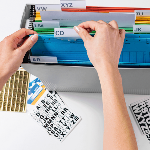 Hände sortieren farbige Ordner in einer Aufbewahrungsbox mit beschrifteten Trennblättern. Daneben liegen Etiketten und Aufkleber.