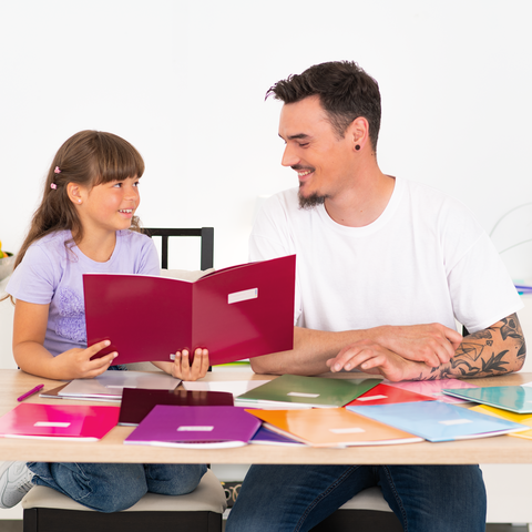 Ein Vater und seine Tochter sitzen an einem Tisch, lächelnd, während sie ein farbiges Heft betrachten. Bunte Hefte liegen vor ihnen.