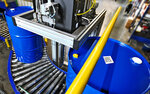 Ein automatisiertes Fördersystem transportiert blaue Fässer in einer Lagerumgebung, während ein Roboterarm über den Fässern positioniert ist.