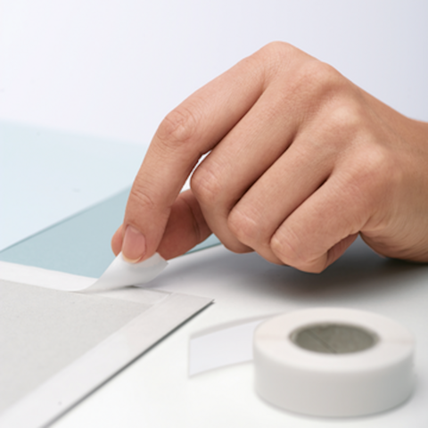 Eine Hand entfernt ein Stück Klebeband von einer grauen Oberfläche, während ein weiteres Stück Klebeband daneben liegt.