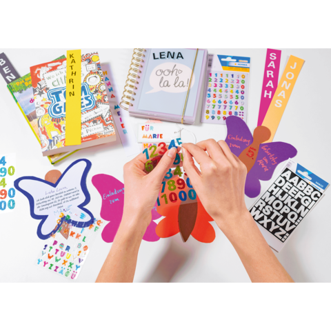 Hände platzieren bunte Aufkleber auf Schmetterlingen und Karten, um kreative Bastelprojekte zu gestalten. Bücher und Sticker sind ebenfalls sichtbar.