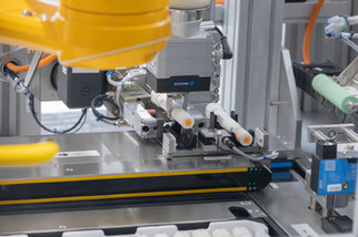 Industrielle Roboterarm mit Greifmechanismus, der Objekte auf einem Förderband bearbeitet. Technische Komponenten und Sensoren sind sichtbar.