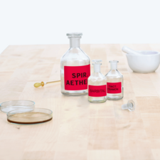 Flaschen mit roten Etiketten, beschriftet mit "Spir. Aether", "Barbital" und "Tinct. Tramon", stehen auf einem Tisch neben einem Mörser.