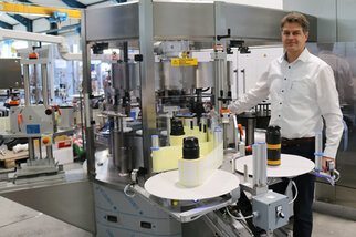 Ein Mann steht neben einer modernen Abfüll- und Etikettiermaschine in einer Produktionshalle, umgeben von Flaschen und Maschinen.