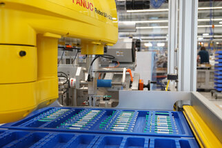 Industrieroboter von FANUC, der Teile aus blauen Behältern greift. Im Hintergrund sind weitere Maschinen und Arbeitsstationen sichtbar.