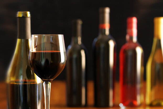 Ein Glas Rotwein steht vor mehreren Weinflaschen in verschiedenen Farben auf einem Tisch.