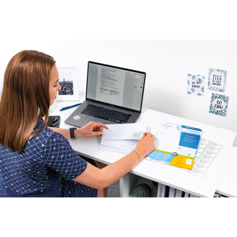 Eine Frau sitzt an einem Schreibtisch, arbeitet am Laptop und sortiert Dokumente und Briefe. Auf dem Tisch liegen verschiedene Unterlagen.