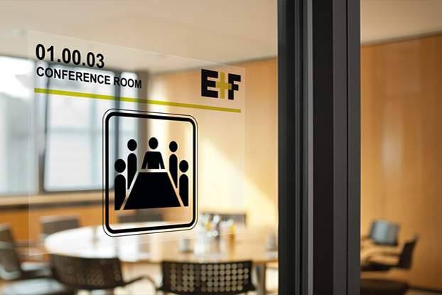 Visitenkarte und Umschlag auf einem Tisch, daneben ein Glastürschild mit einem Konferenzraum-Symbol und drei Parfümflaschen.