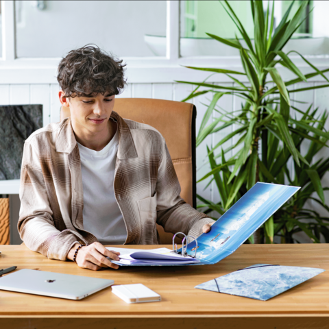 Ein junger Mann sitzt an einem Schreibtisch, schaut auf einen Ordner und arbeitet an einem Laptop. Pflanzen sind im Hintergrund sichtbar.