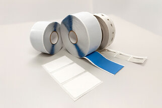Zwei Rollen Etikettenmaterial, eine mit blauer und eine mit weißer Oberfläche, sowie mehrere leere Etiketten auf einem Tisch.