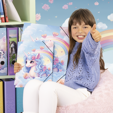 Ein Mädchen in einem blauen Pullover hält zwei Ordner mit einem Einhorn-Motiv und lächelt vor einem bunten Hintergrund.