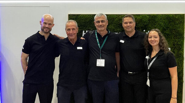 Fünf Personen in schwarzer Kleidung stehen nebeneinander vor einer Wand mit dem HERMA-Logo und dem Slogan „Coating your success“.