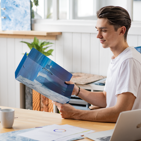 Ein junger Mann sitzt an einem Tisch und betrachtet eine Mappe mit einer Landschaftsaufnahme, während er eine Tasse Kaffee hält.