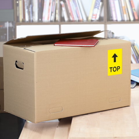 Kartonbox mit einem roten Heft auf der Oberseite, beschriftet mit einem gelben "TOP"-Schild, steht auf einem Tisch in einer Bibliothek.