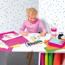 Ein kleines Mädchen sitzt an einem Tisch und malt mit Wasserfarben. Um sie herum liegen bunte Zeichnungen und Bastelmaterialien.
