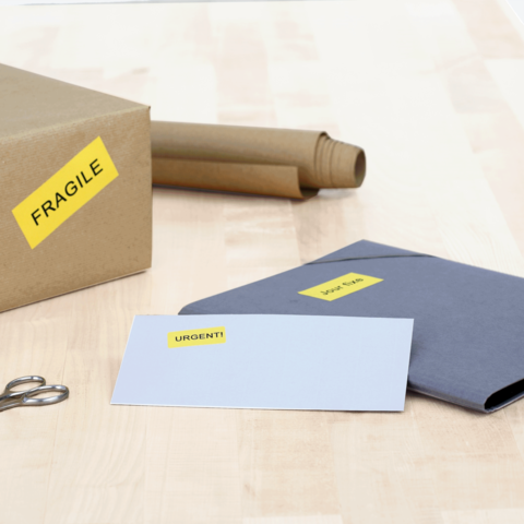 Ein Karton mit dem Aufkleber "FRAGILE", daneben ein graues Kuvert mit "URGEN!T" und ein weißes Blatt. Scheren liegen bereit.